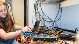 A student works on a complex setup of electronic components and wires arranged on a desk in a technical lab environment.