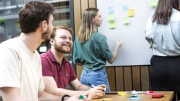 Computer Science students collaborate on a project, with one writing on a sticky-note-covered whiteboard and others discussing.