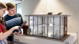 Two Electrical Engineering students examine a detailed architectural model of a modern building, with one using a tablet.