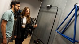 Two Communication Networks students examine a wall-mounted antenna, with one pointing at it during a discussion.