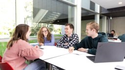 Four mathematics students are sitting together at a table in a modern study space, engaged in discussion.