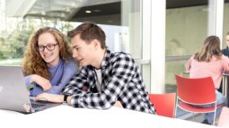 Two math students work on a laptop in a bright, modern space with large windows and others visible in the background.